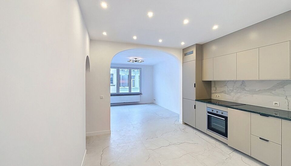 Bij het betreden van dit volledig gerenoveerde appartement wordt u verwelkomd in een stijlvolle inkomhal met een elegante, afgeronde deuropening.
Direct aan de inkom bevindt zich een bureauruimte met parketlaminaat en een gloednieuw raam, ideaal voor thui