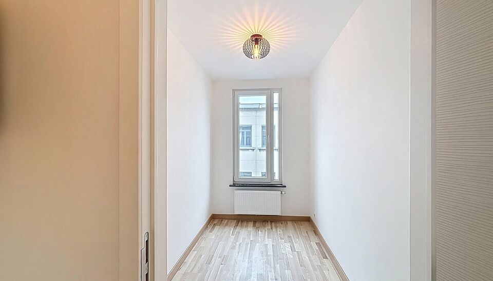 Bij het betreden van dit volledig gerenoveerde appartement wordt u verwelkomd in een stijlvolle inkomhal met een elegante, afgeronde deuropening.
Direct aan de inkom bevindt zich een bureauruimte met parketlaminaat en een gloednieuw raam, ideaal voor thui